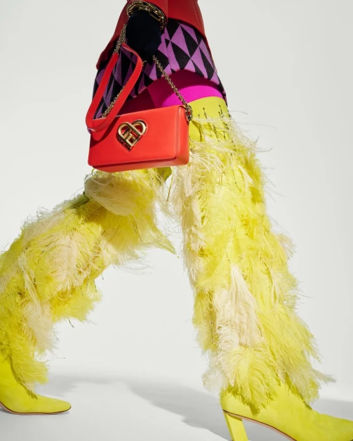 Fashion editorial featuring yellow feather-embellished pants and boots paired with a bright red heart-shaped handbag on a white background.