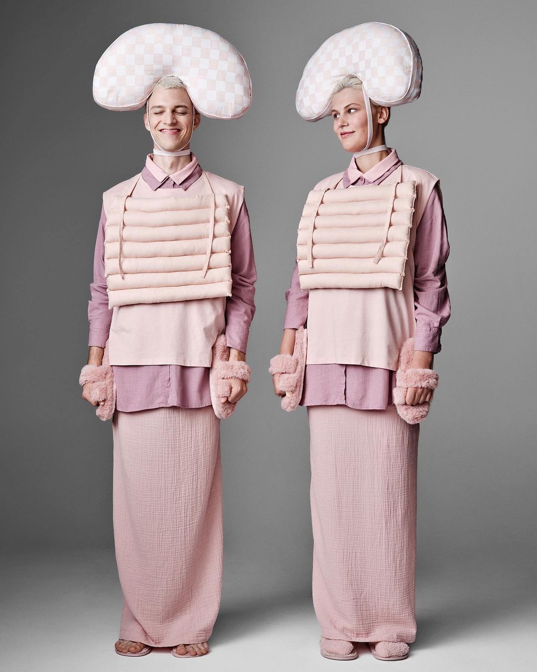 Two people stand side-by-side in pink and white clothing with padded headgear and arm coverings, against a grey studio backdrop.