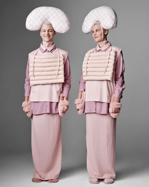Two people stand side-by-side in pink and white clothing with padded headgear and arm coverings, against a grey studio backdrop.