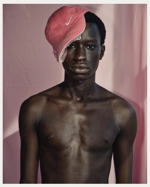 Portrait of a shirtless Black man wearing a pink mesh Nike cap, set against a light pink backdrop, in a studio photography style.