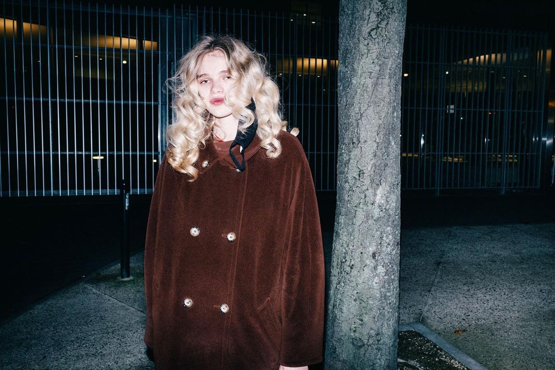 Portrait of a young adult female with wavy blonde hair, wearing a brown double-breasted coat, standing outdoors by a tree at night.