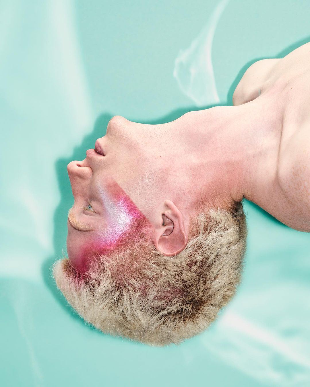 Upside-down portrait of a male with makeup, light skin, and short blonde hair, on a blue backdrop with soft, diffused light.