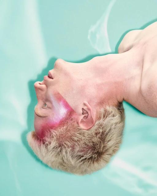 Upside-down portrait of a male with makeup, light skin, and short blonde hair, on a blue backdrop with soft, diffused light.