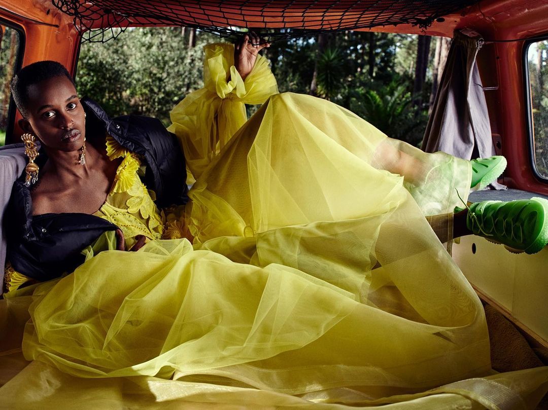 Adult woman in a yellow dress and black jacket, lying in the back of an orange van surrounded by green foliage. Fashion shot.