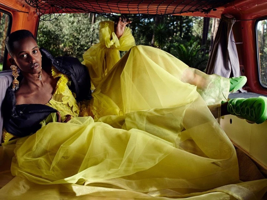 Adult woman in a yellow dress and black jacket, lying in the back of an orange van surrounded by green foliage. Fashion shot.