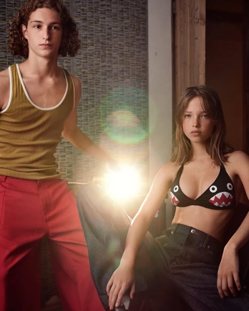 Two young adults pose indoors; one stands in a yellow tank top and red pants, while the other sits wearing a graphic top and jeans.