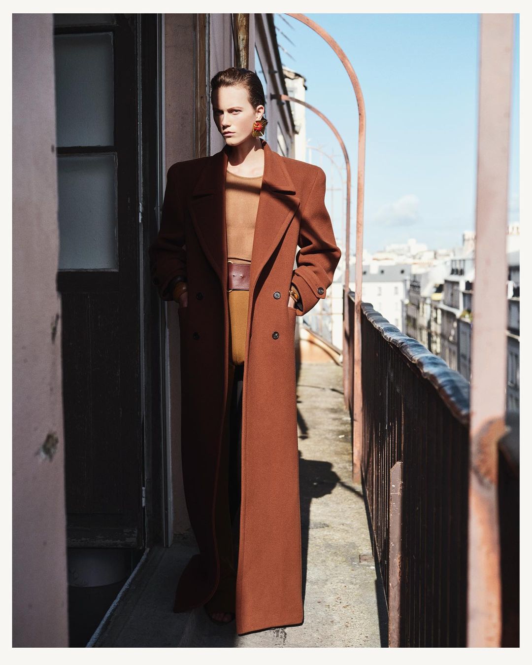 Model wearing a long brown coat standing on a Parisian balcony overlooking the cityscape in bright daylight
