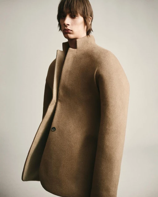 A young adult male with brown hair poses in a studio wearing a beige, oversized coat. The background is a neutral grey colour.