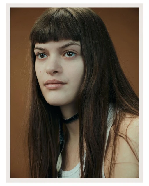 Portrait of a young woman with long brown hair and bangs wearing a white tank top against a solid brown backdrop in a photo studio.