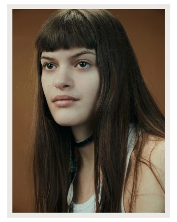 Portrait of a young woman with long brown hair and bangs wearing a white tank top against a solid brown backdrop in a photo studio.