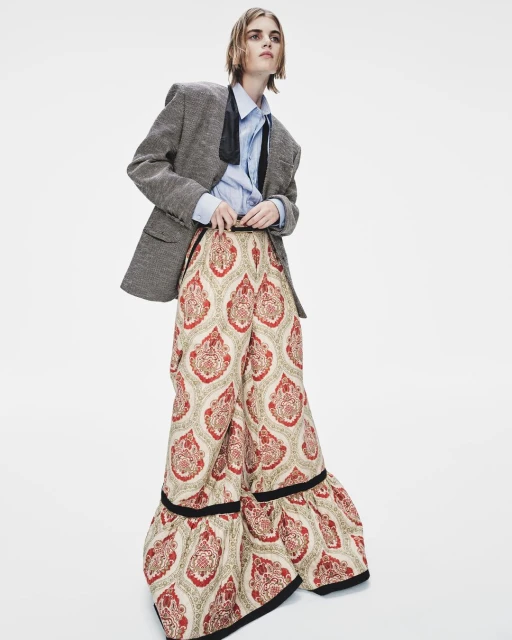 Fashion photo featuring a slender person in a light blue shirt, patterned palazzo pants, and a tweed blazer, posing against a white backdrop.