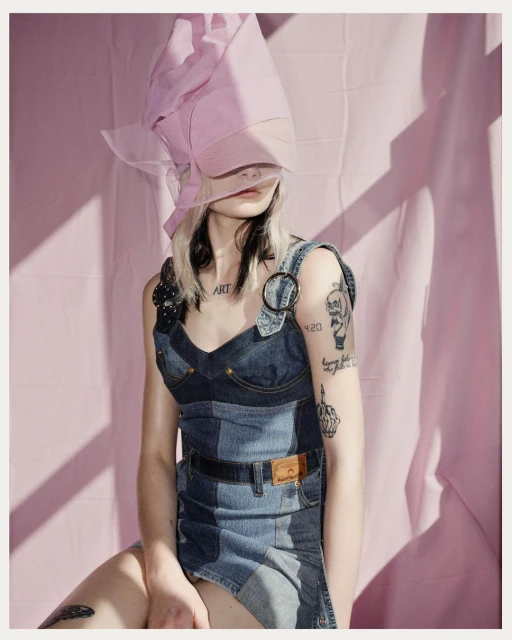 A fashion portrait features a stylish adult female with tattoos, wearing a denim dress and a pink head covering, set against a pink backdrop.