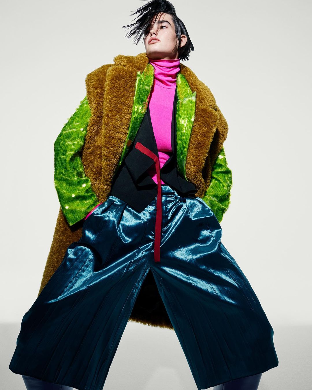 Fashion model in statement outerwear: a textured brown coat, a shimmering green jacket, and wide-leg blue pants in a studio shot.
