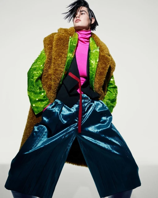 Fashion model in statement outerwear: a textured brown coat, a shimmering green jacket, and wide-leg blue pants in a studio shot.