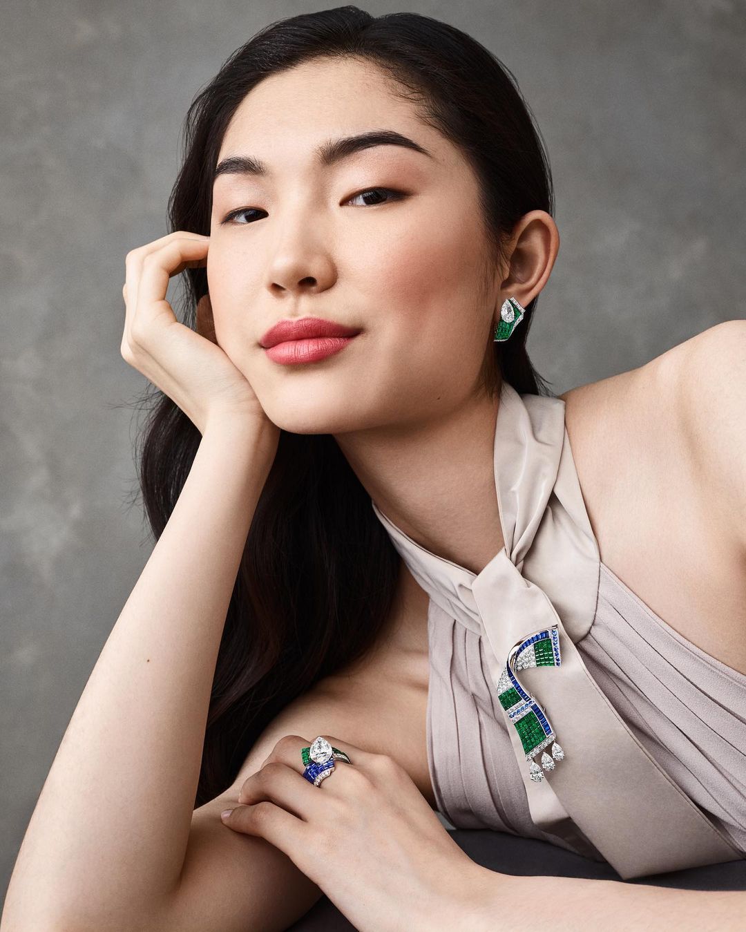 Asian woman poses with jewelry. She has on emerald earrings and a ring and also a tie-like jewelry piece around her neck.