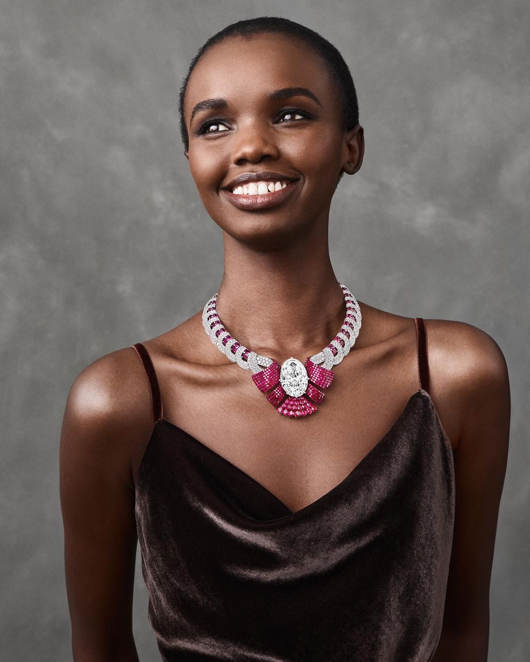 Elegant Black woman wearing a bejeweled necklace with rubies and diamonds. She is smiling and looking off to the side with a grey background.