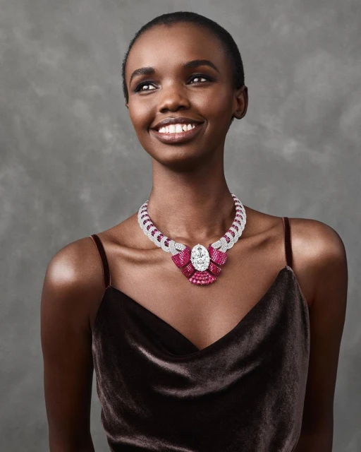 Elegant Black woman wearing a bejeweled necklace with rubies and diamonds. She is smiling and looking off to the side with a grey background.