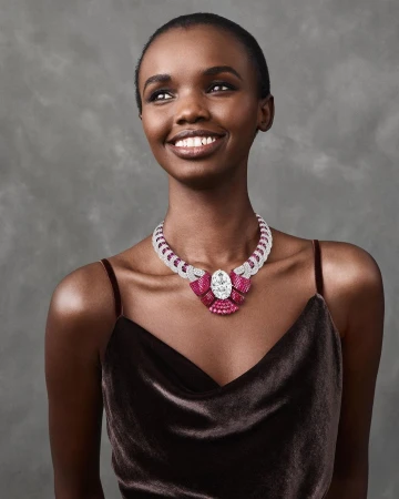 Elegant Black woman wearing a bejeweled necklace with rubies and diamonds. She is smiling and looking off to the side with a grey background.