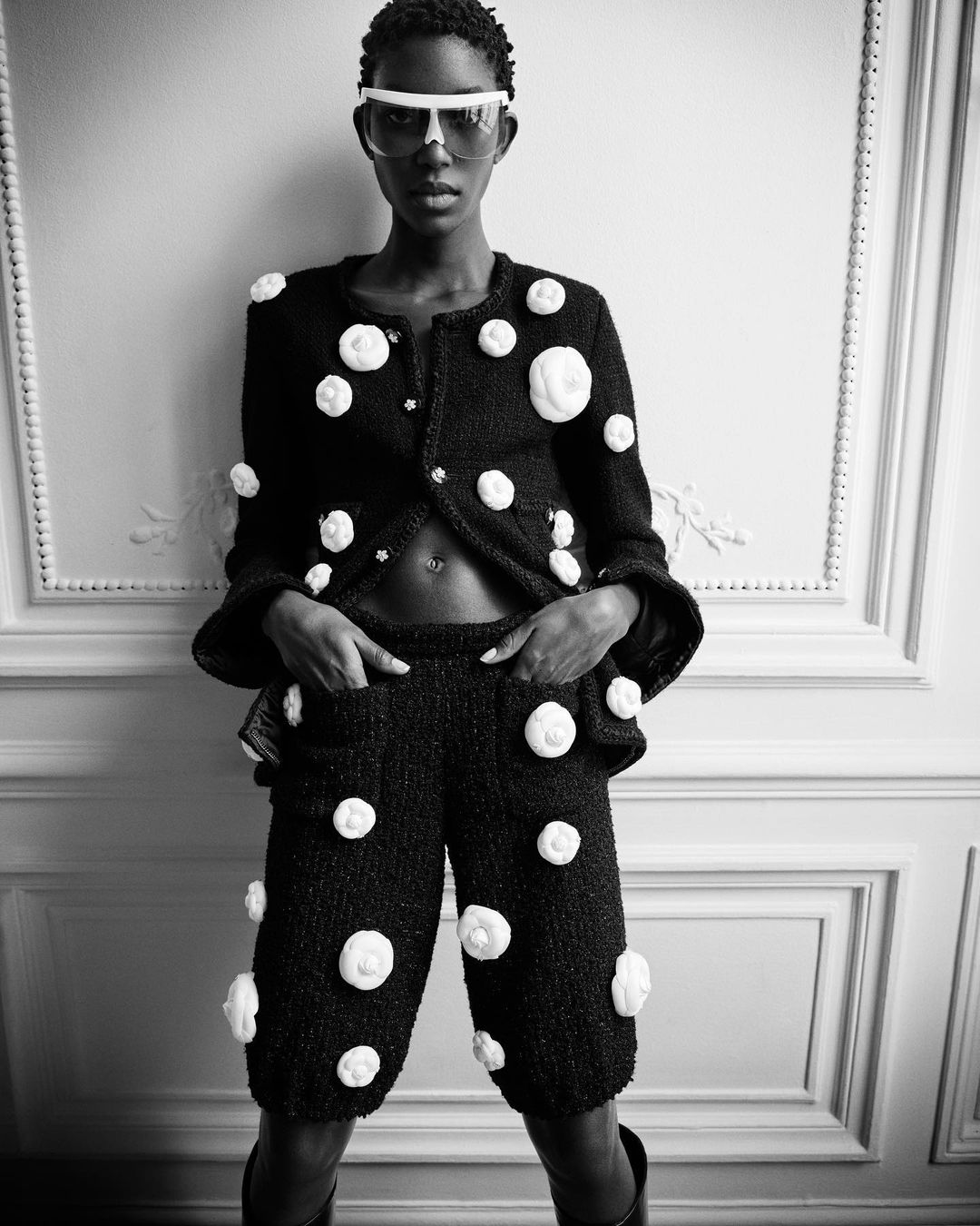 Black and white fashion portrait of a Black woman wearing a jacket and shorts adorned with white flower embellishments against a wall.