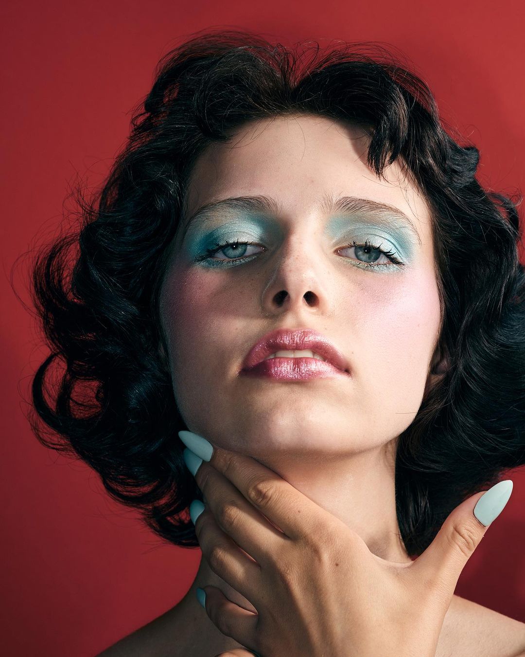 Close-up portrait of a woman with short dark hair, blue eyeshadow, and light blue manicured nails held near her neck against a red background