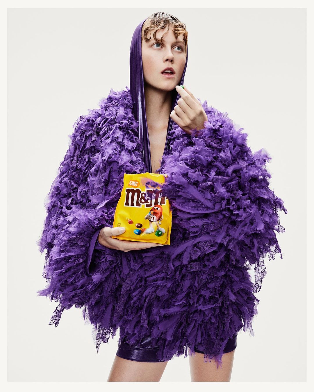 Adult woman in purple feathered outfit poses in studio with M&M's bag and candy. Fashion and food photography. Bright, colorful studio shot.