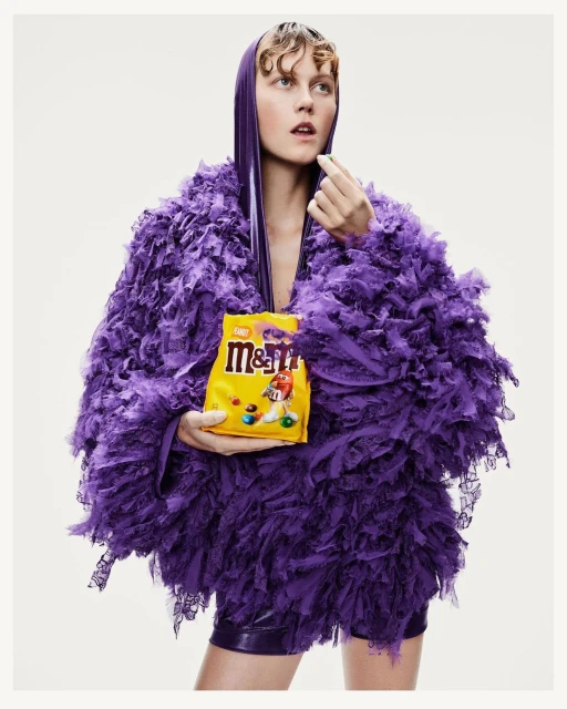 Adult woman in purple feathered outfit poses in studio with M&M's bag and candy. Fashion and food photography. Bright, colorful studio shot.