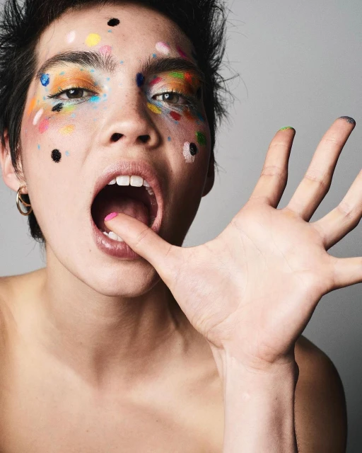Close-up portrait of a fair-skinned adult female with colorful makeup touching her mouth with a pink-painted fingertip.