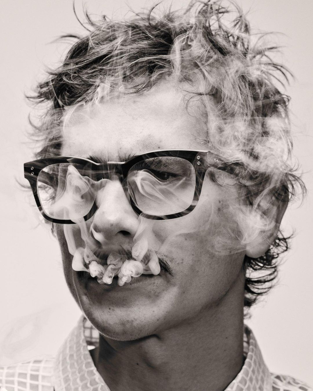 Monochrome portrait of a man with glasses, mustache, and smoke around the face, set against a plain background in a studio.