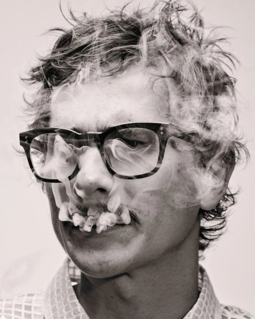 Monochrome portrait of a man with glasses, mustache, and smoke around the face, set against a plain background in a studio.