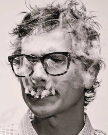 Monochrome portrait of a man with glasses, mustache, and smoke around the face, set against a plain background in a studio.