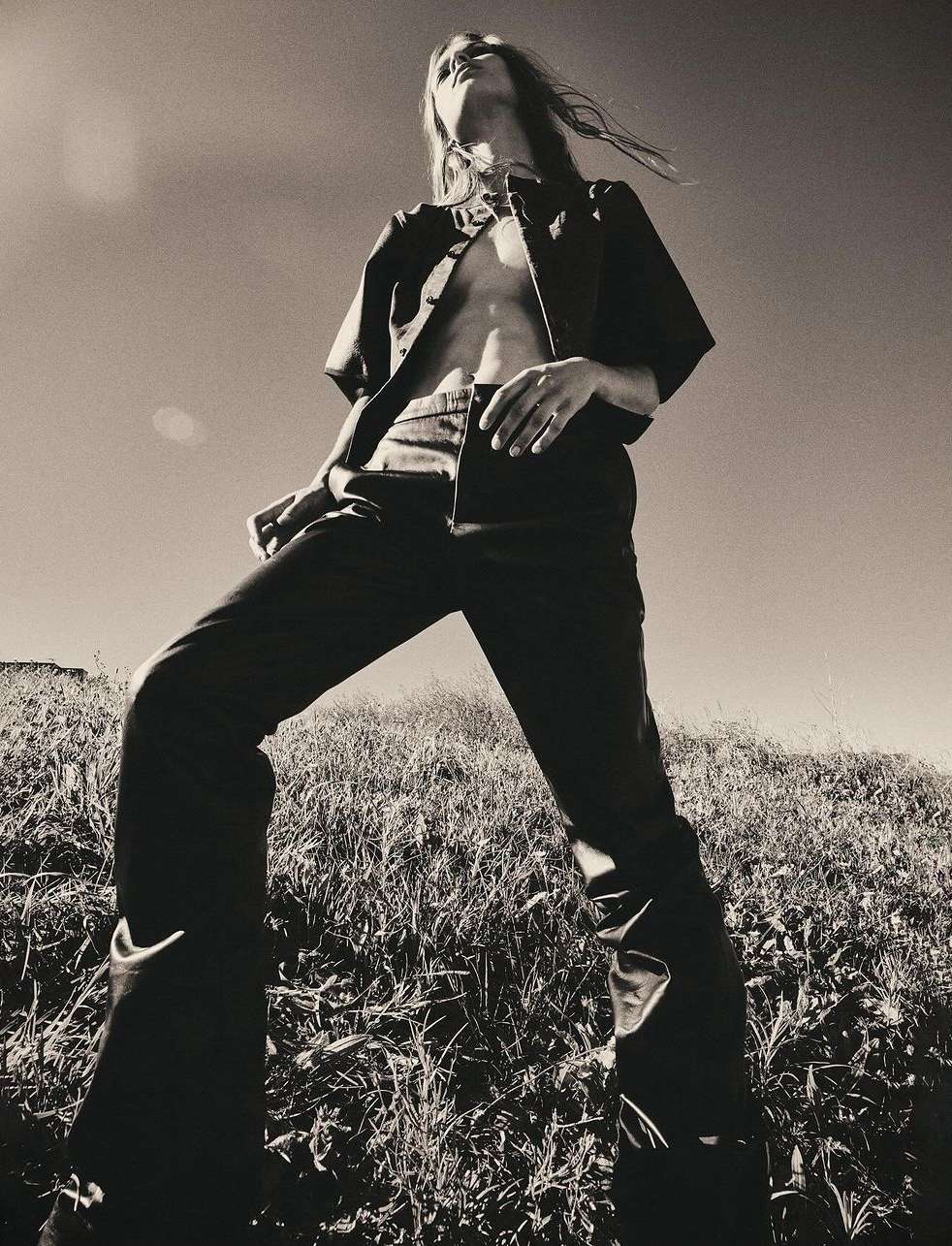 A low-angle black and white shot of a person with long hair wearing dark, partially unbuttoned shirt and pants standing on a grassy hill under a clear sky.