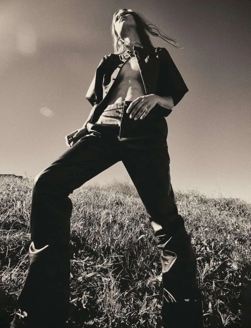 A low-angle black and white shot of a person with long hair wearing dark, partially unbuttoned shirt and pants standing on a grassy hill under a clear sky.