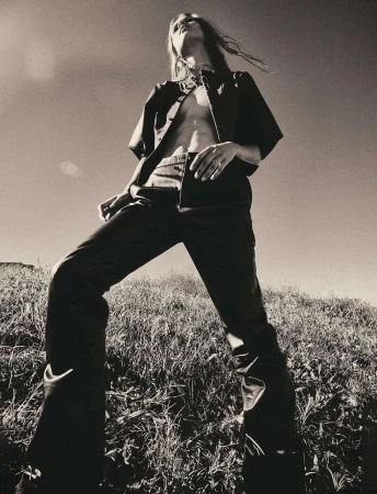 A low-angle black and white shot of a person with long hair wearing dark, partially unbuttoned shirt and pants standing on a grassy hill under a clear sky.