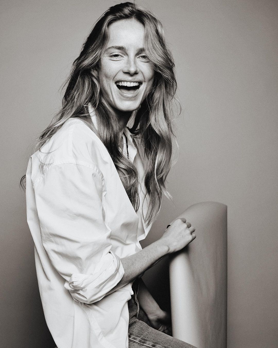 Portrait of a laughing woman with long hair wearing an unbuttoned white shirt against a neutral grey studio backdrop.