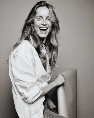 Portrait of a laughing woman with long hair wearing an unbuttoned white shirt against a neutral grey studio backdrop.