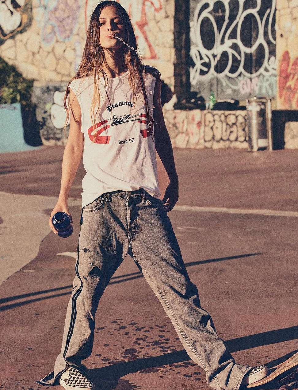 Young adult poses with drink on skateboard in an urban skatepark. Graffiti wall backdrop and overcast light enhance the casual scene.