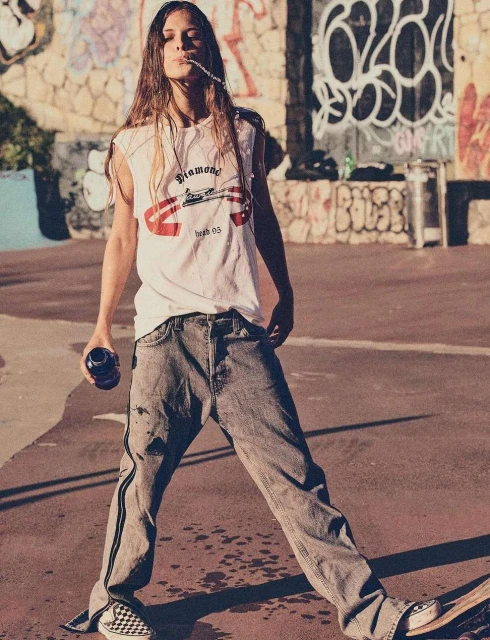 Young adult poses with drink on skateboard in an urban skatepark. Graffiti wall backdrop and overcast light enhance the casual scene.