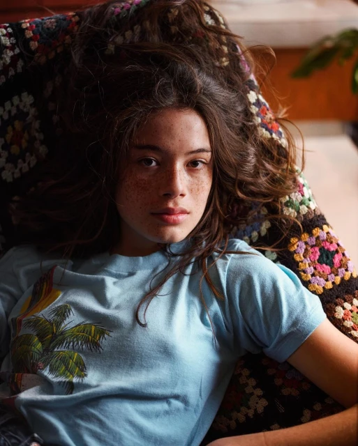 Portrait of a young adult woman laying on a patterned blanket, wearing a blue t-shirt with a palm tree graphic.