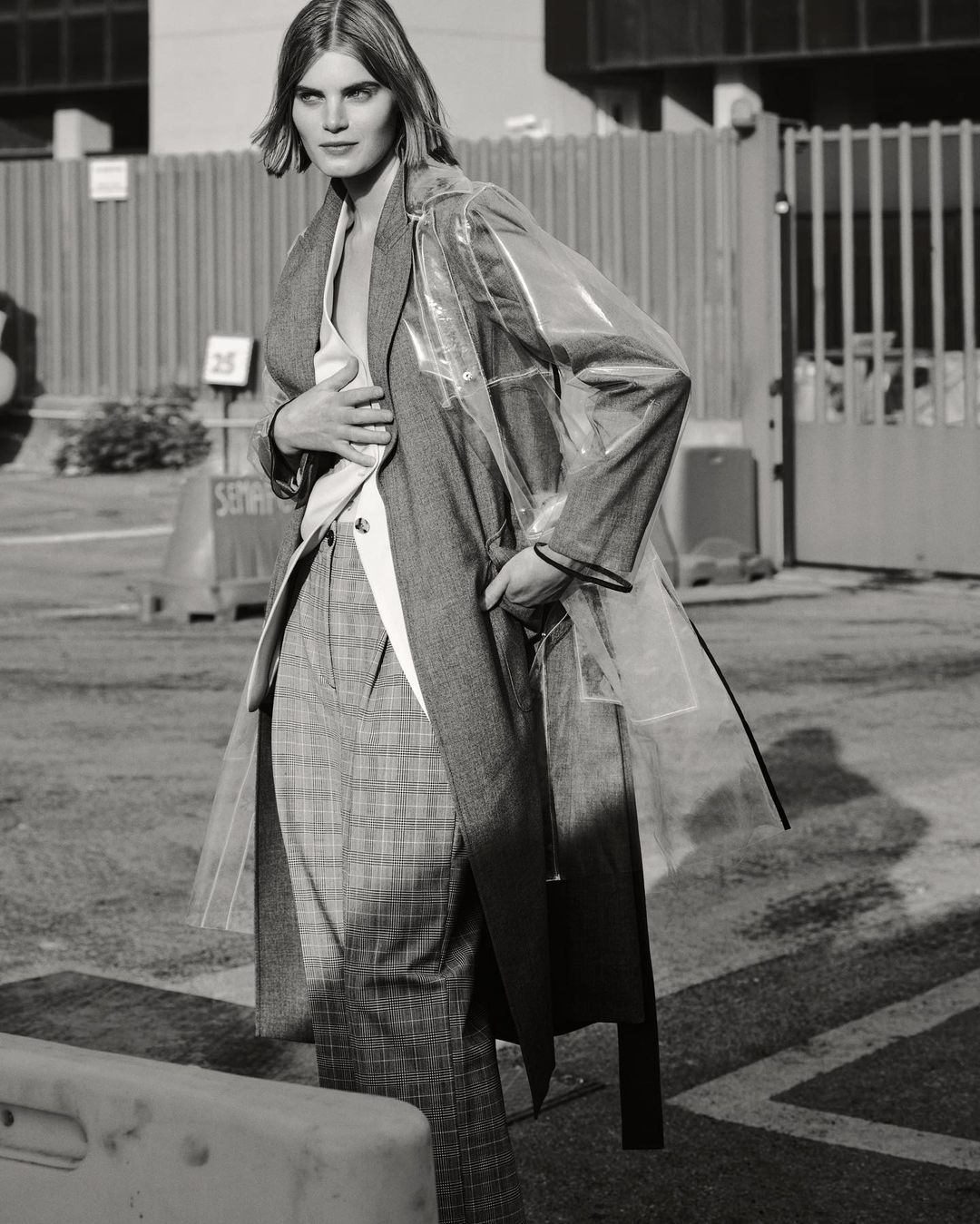 Monochrome portrait of a woman in a clear coat, grey blazer, and plaid pants, standing in an urban environment. Street style fashion shot.
