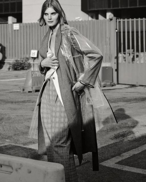 Monochrome portrait of a woman in a clear coat, grey blazer, and plaid pants, standing in an urban environment. Street style fashion shot.