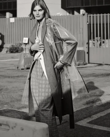 Monochrome portrait of a woman in a clear coat, grey blazer, and plaid pants, standing in an urban environment. Street style fashion shot.