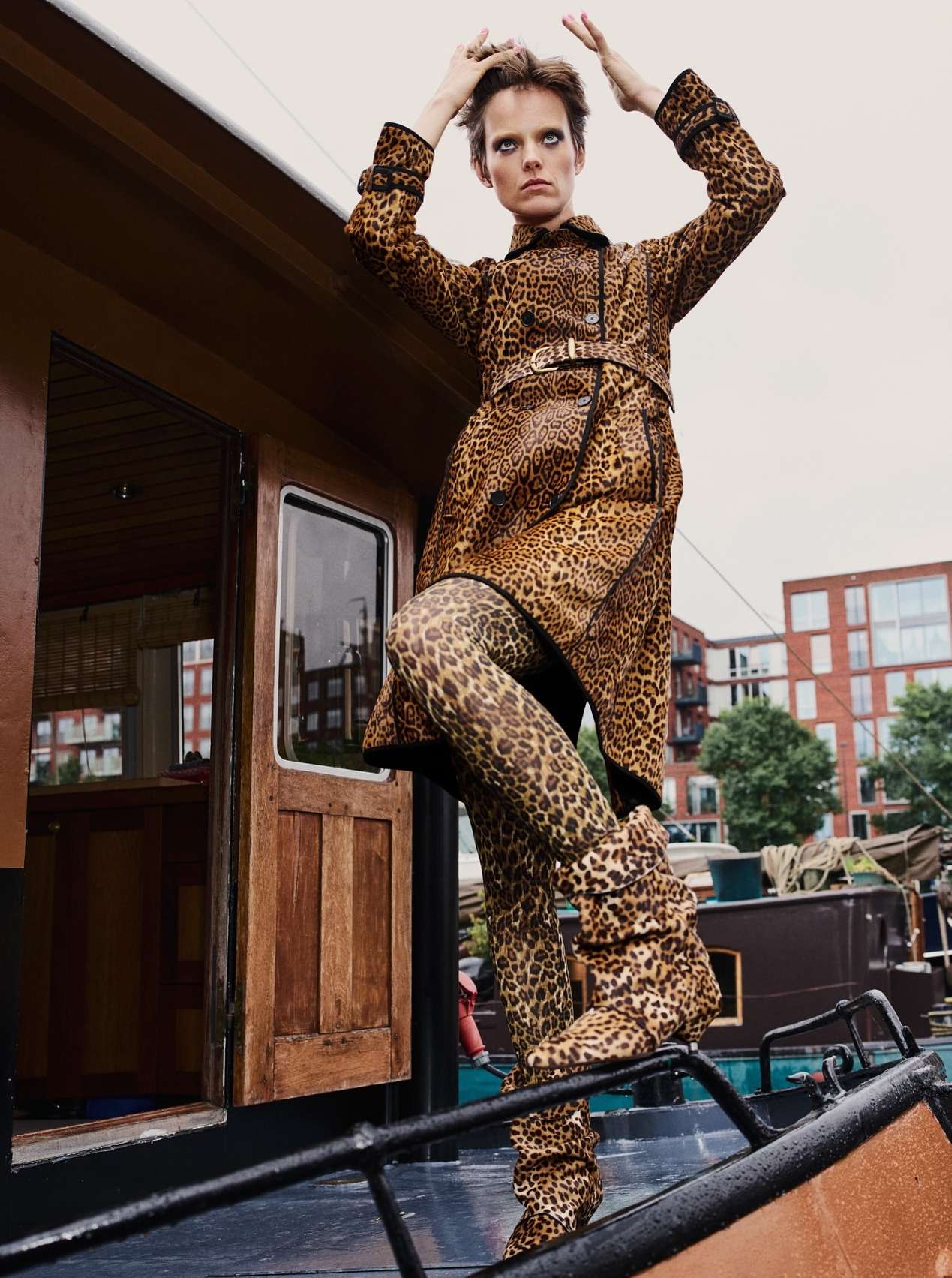 Fashionable woman in leopard print coat and leggings poses on boat against city buildings. Bold, stylish outdoor fashion shot.