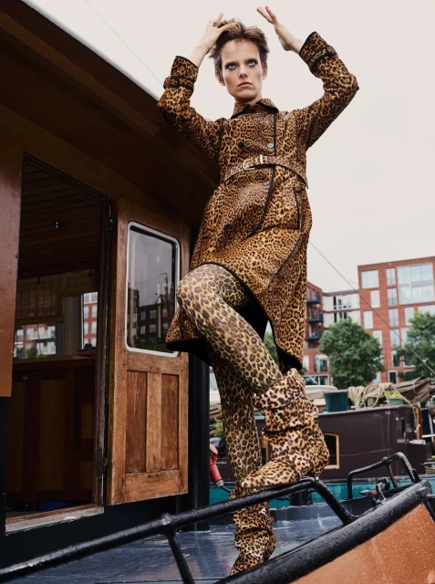 Fashionable woman in leopard print coat and leggings poses on boat against city buildings. Bold, stylish outdoor fashion shot.