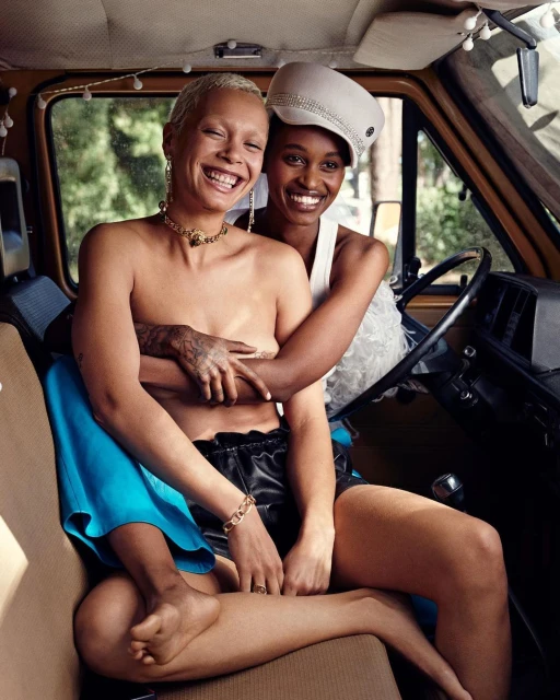 Two women embrace and smile inside a vintage van with string lights; one is topless with a gold necklace and the other wears a white hat.