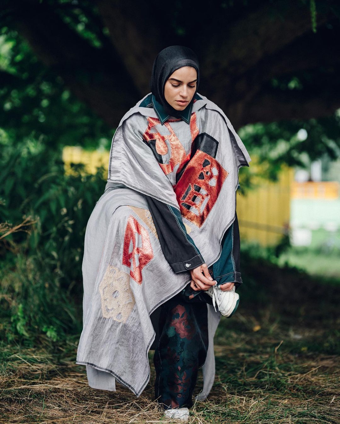 Woman in hijab and avant-garde fashion poses in an outdoor setting with lush green foliage and muted lighting, creating a stylish portrait.