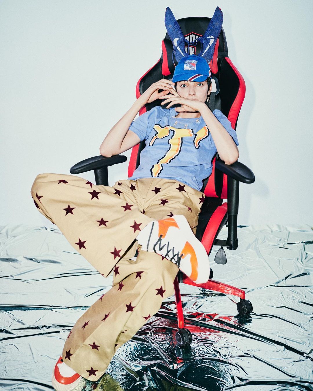 Young adult poses in a gamer chair, wearing star-patterned pants, a t-shirt, baseball cap, and sneakers against a shiny backdrop.
