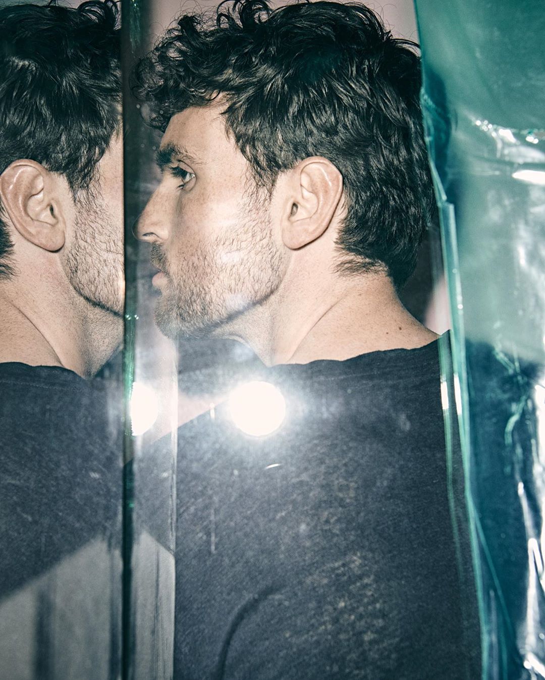 Profile portrait of a man with dark hair reflected in glass, wearing a black shirt, against a background with blue plastic wrap.