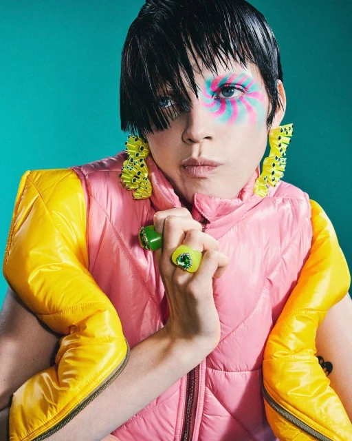 Stylish portrait of a person with colorful spiral face makeup, yellow and pink padded vest, big earrings, and green rings against teal.