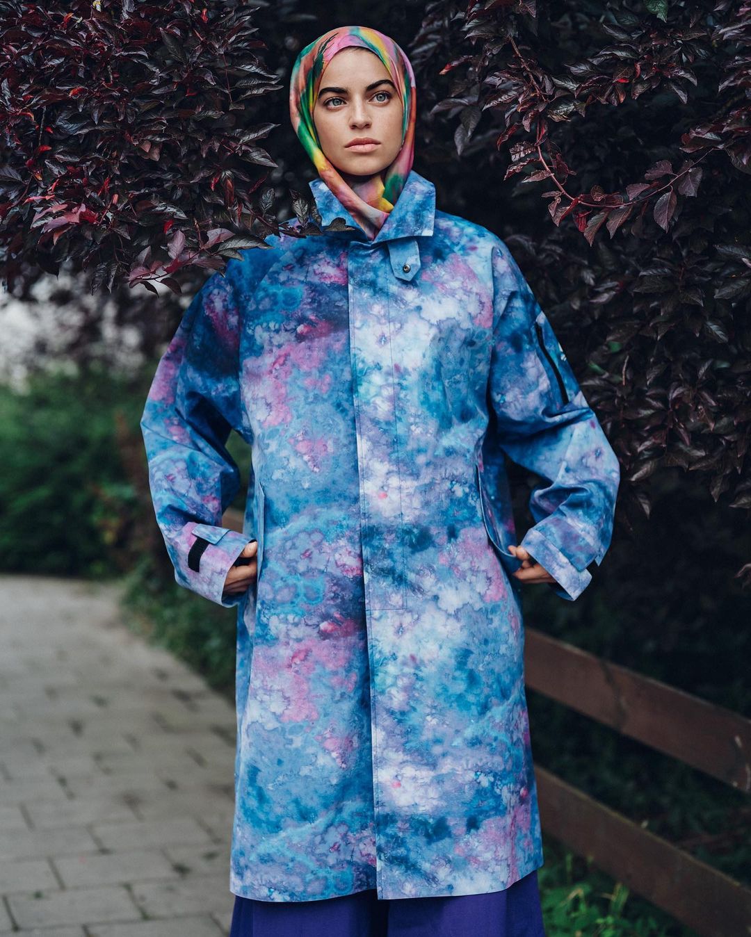 Woman wearing a colorful hijab and a blue and purple patterned coat stands outside against a backdrop of dark foliage and a wooden fence.
