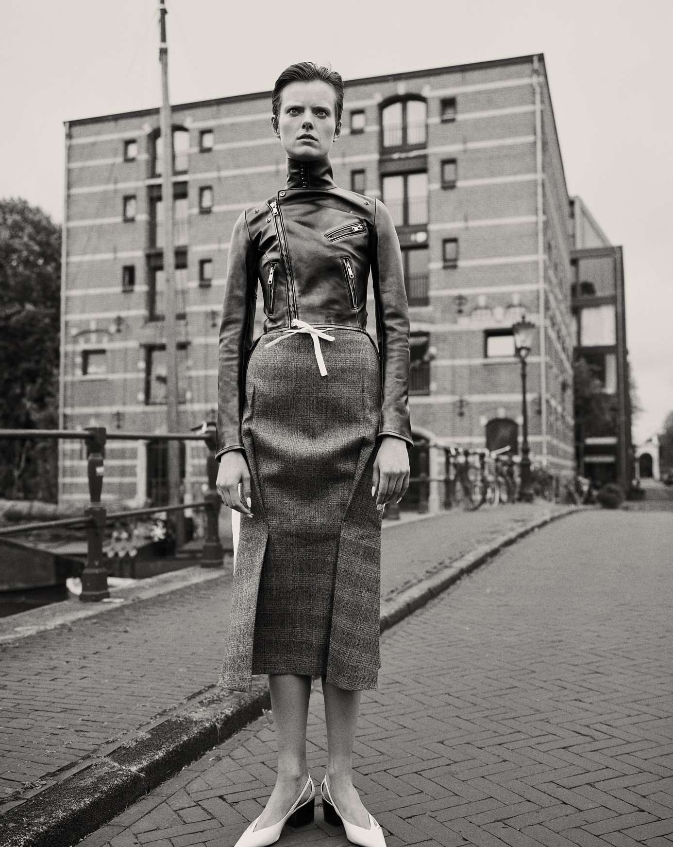 A woman in a leather jacket and skirt poses on a cobblestone street in front of a brick building in a black and white fashion photograph.